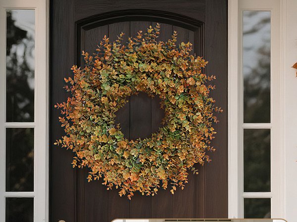 Artificial boxwood fall wreath with green and orange leaves hanging on a dark brown front door with white trim.
