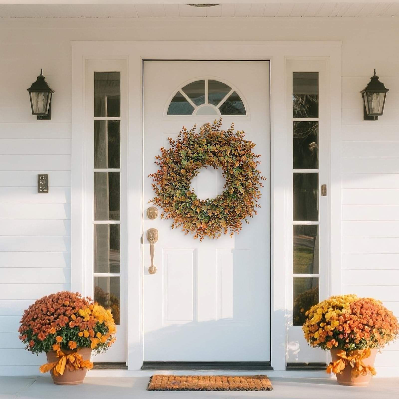 Artificial boxwood fall wreath with green and brown leaves hanging on a white front door with two potted mum plants flanking the entrance.