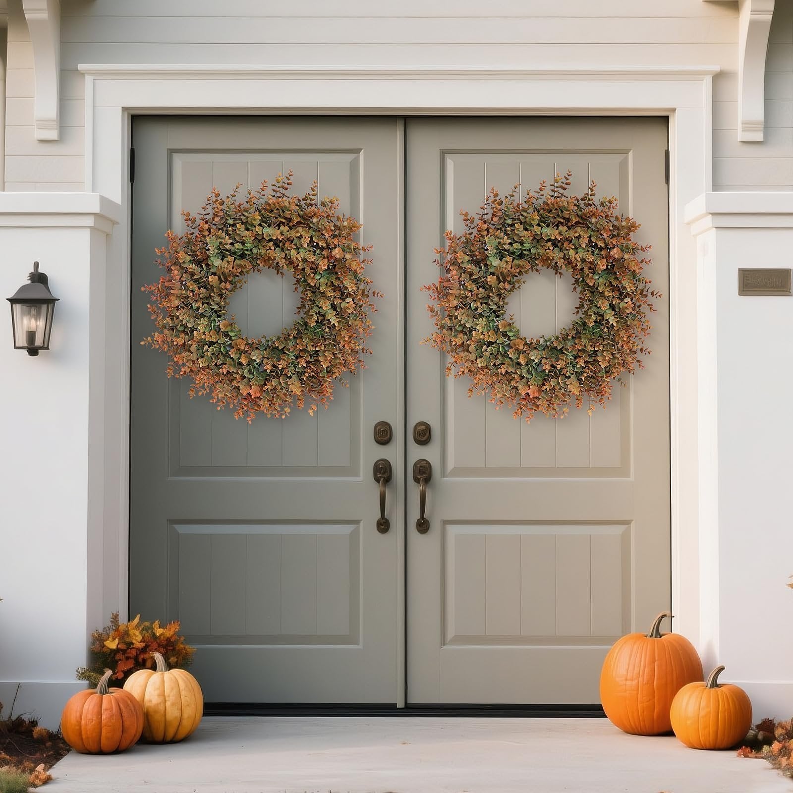 Two artificial boxwood fall wreaths with green and brown leaves hang on double doors, with pumpkins and fall foliage on the porch below.