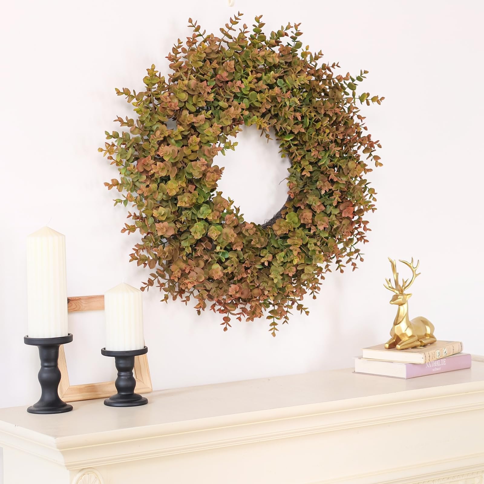 Artificial fall wreath with green and brown leaves hanging on a white wall above a mantelpiece with decor