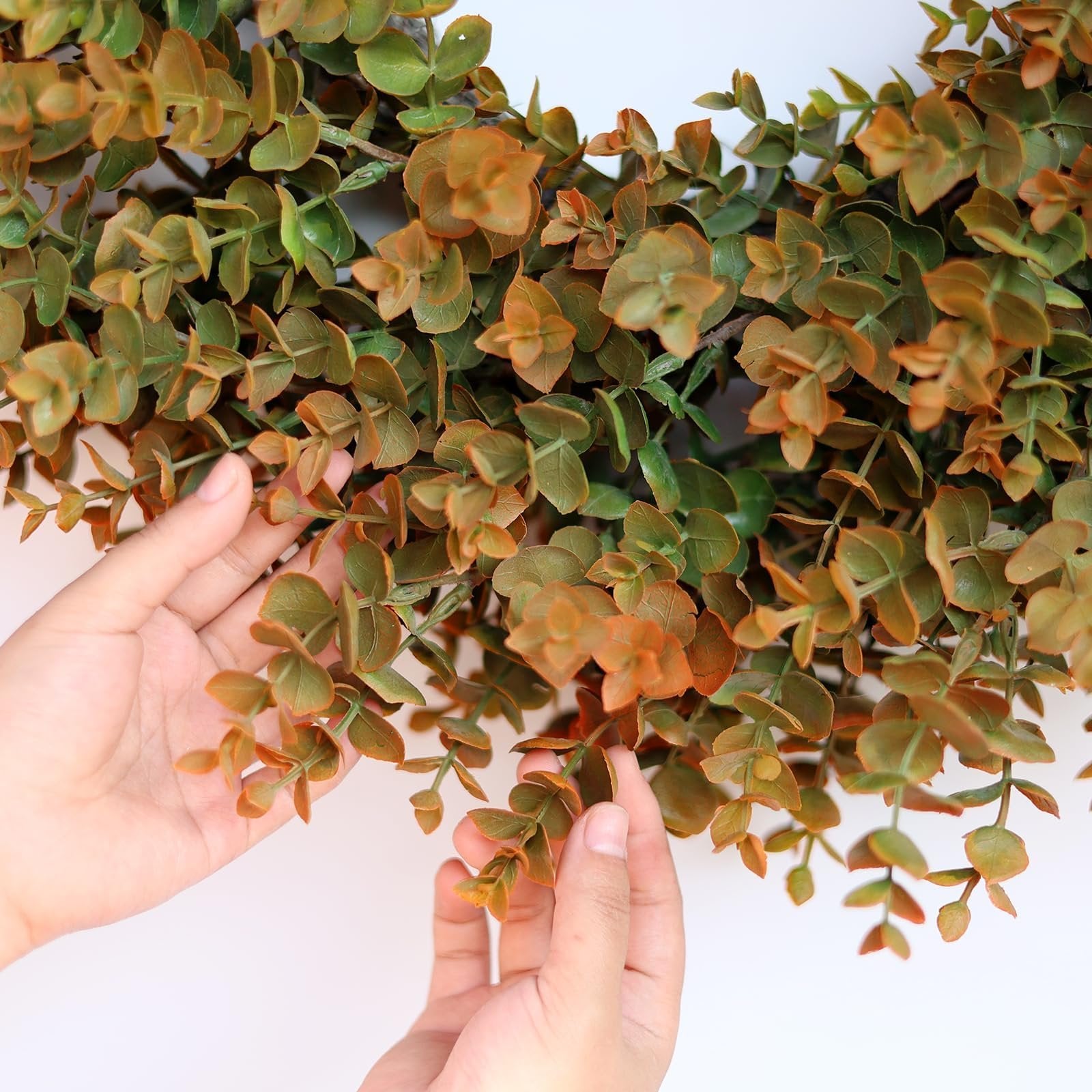 Close-up of hands touching an artificial boxwood wreath with green and brown leaves against a white background.