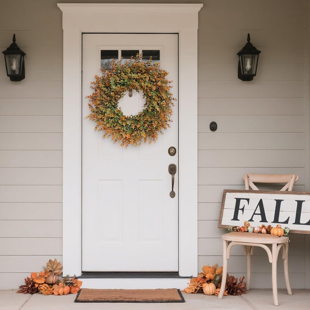 Artificial boxwood fall wreath with green and orange leaves hanging on a white front door, flanked by lanterns and decorated with pumpkins and a 'FALL' sign.