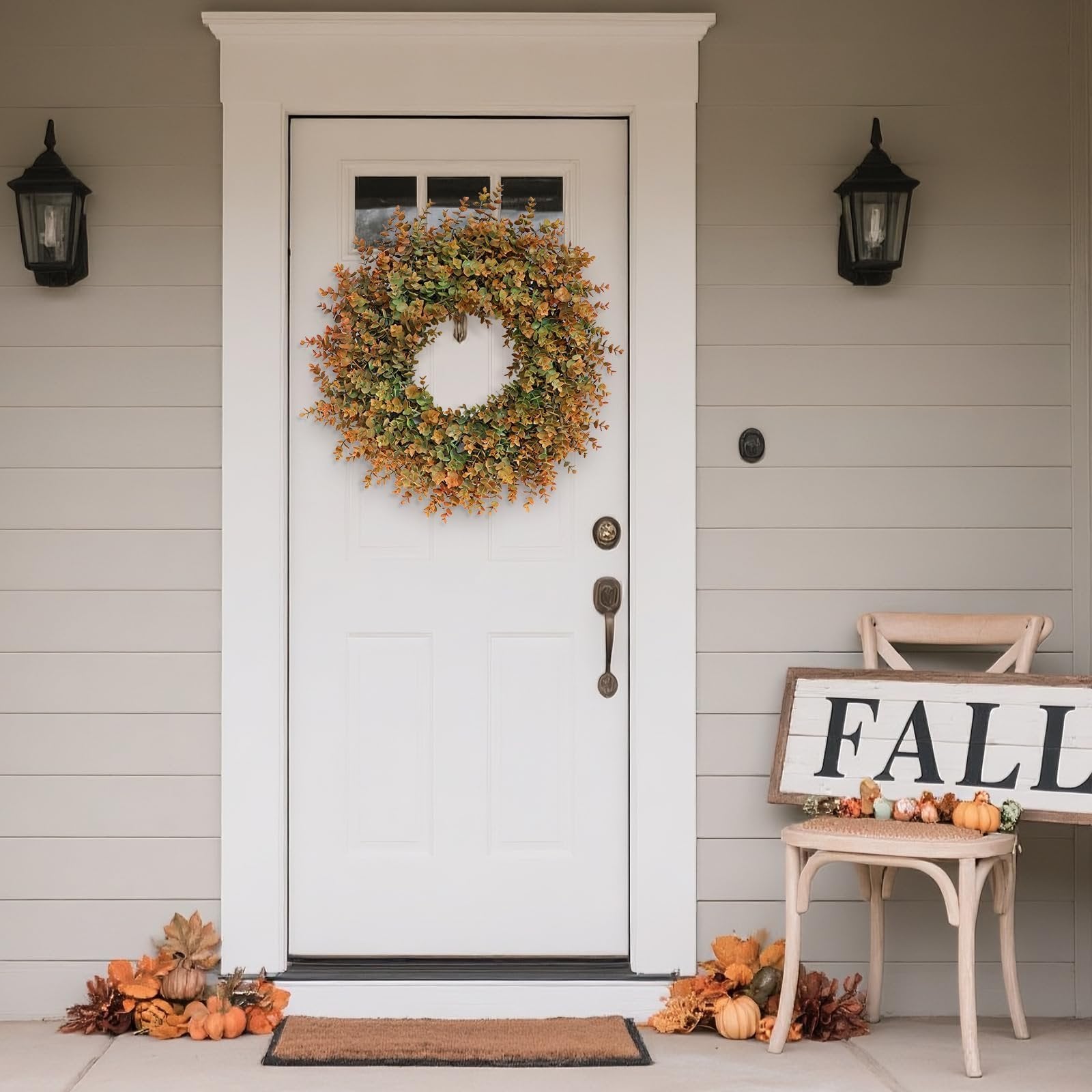Artificial boxwood fall wreath with green and orange leaves hanging on a white front door, flanked by lanterns and decorated with pumpkins and a 'FALL' sign.