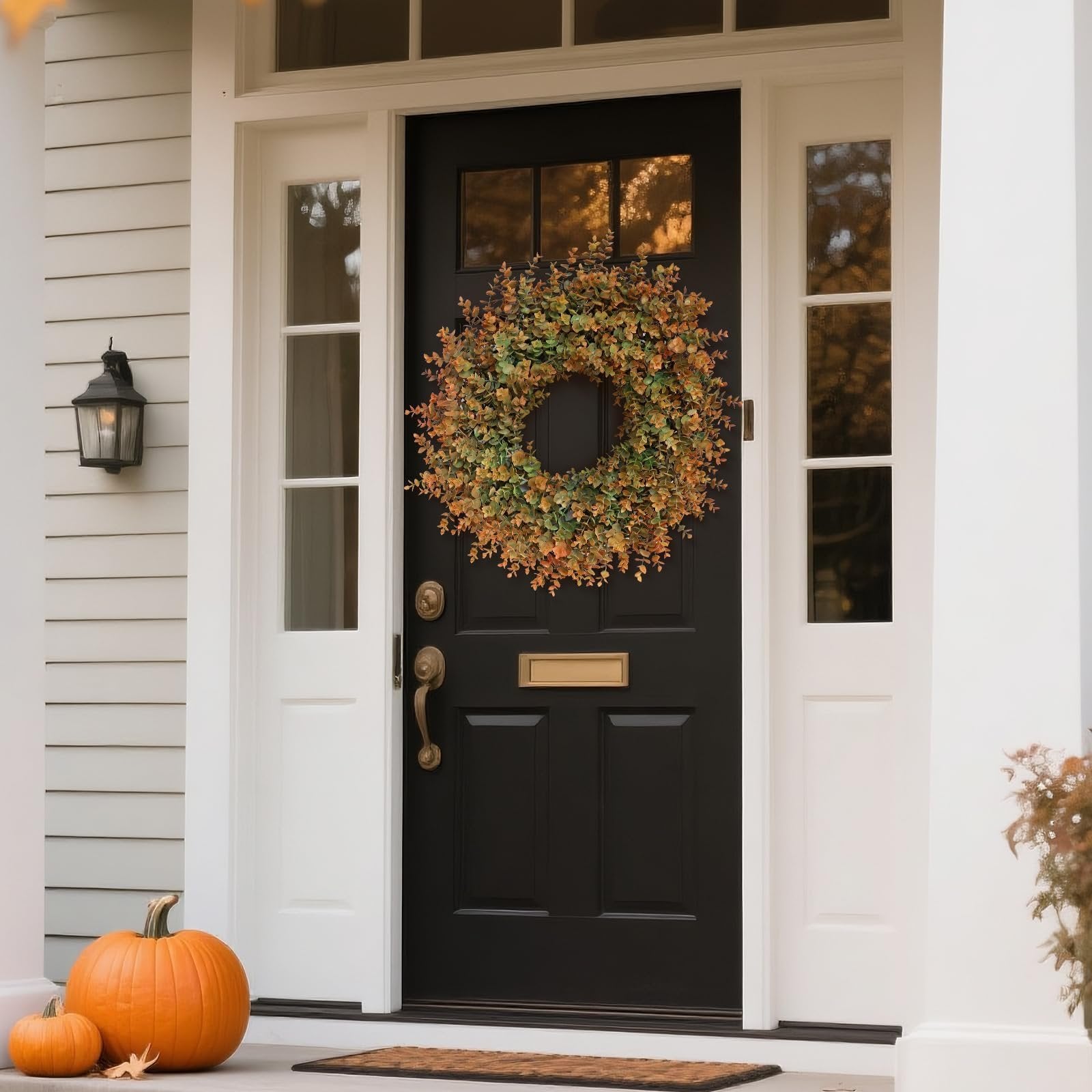 Artificial boxwood fall wreath with green and orange leaves hanging on a black front door with pumpkins on the porch.