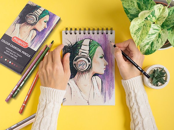 Hands drawing a portrait with colored charcoal pencils, showing pencil box and a notebook sketch.