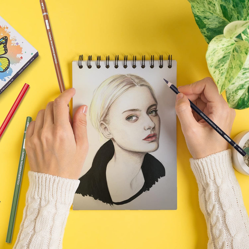Hands drawing a portrait of a woman with charcoal pencils on a sketchbook, surrounded by pencils and a butterfly-themed box.