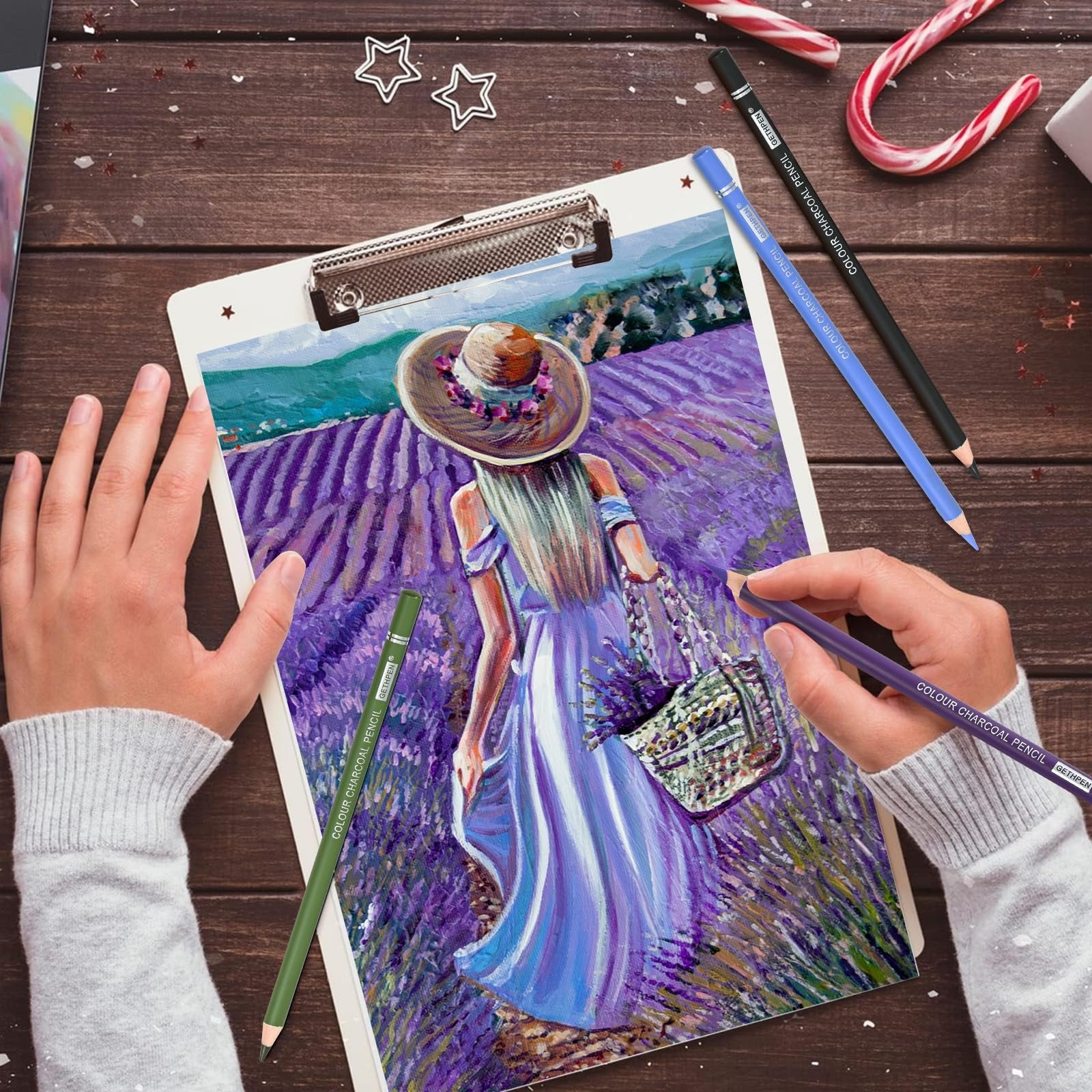 Hands holding charcoal pencils drawing a lavender field scene on a clipboard