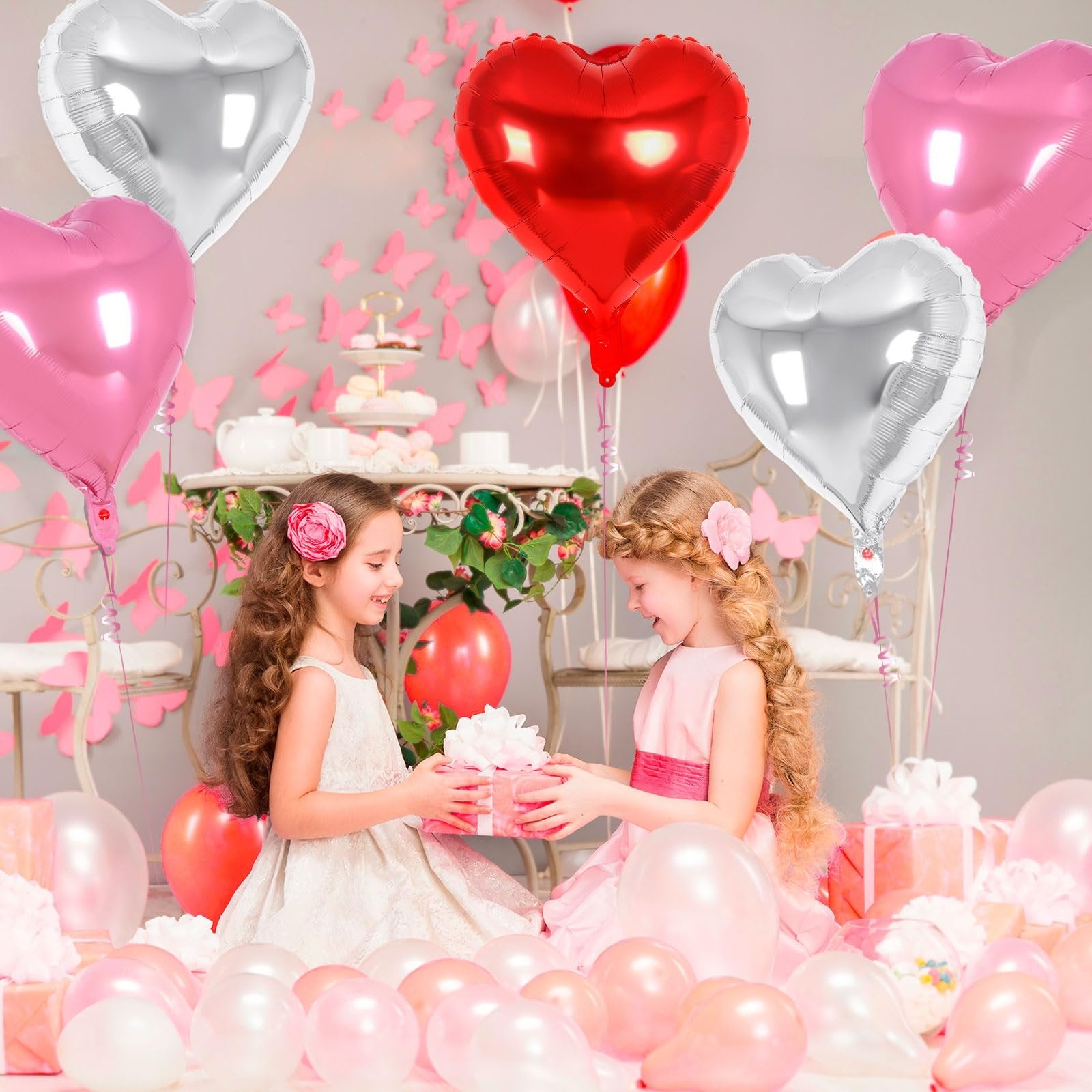Two young girls in party dresses exchanging a gift surrounded by heart-shaped balloons and presents.