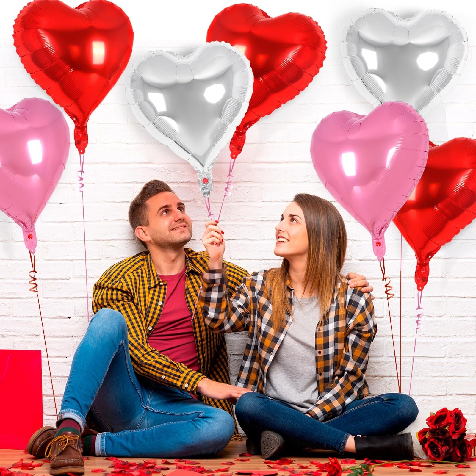 Couple sitting on floor with red, pink, and silver heart foil balloons against a white brick wall