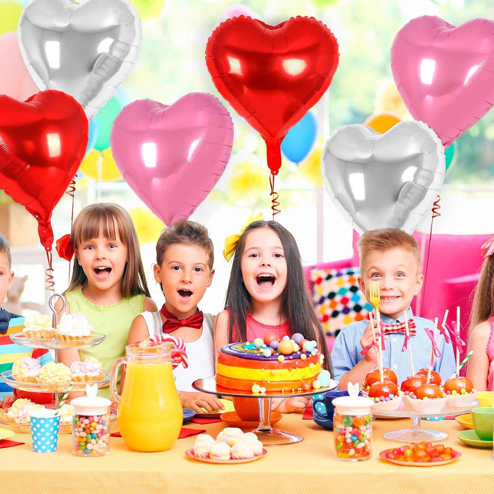 Children at a party table with heart-shaped balloons, cake, and candy.