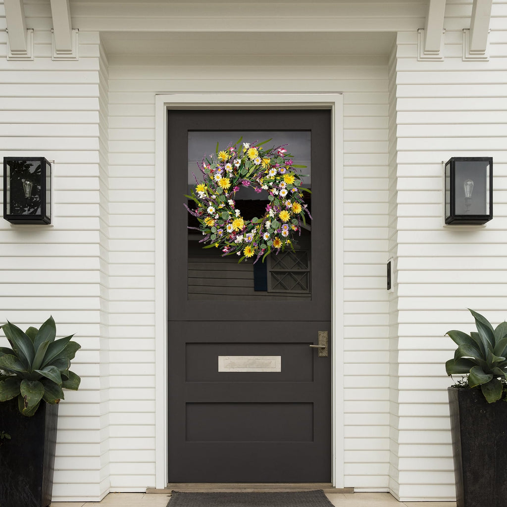 Artificial daisy and lavender wildflower wreath hanging on a dark front door with white siding.