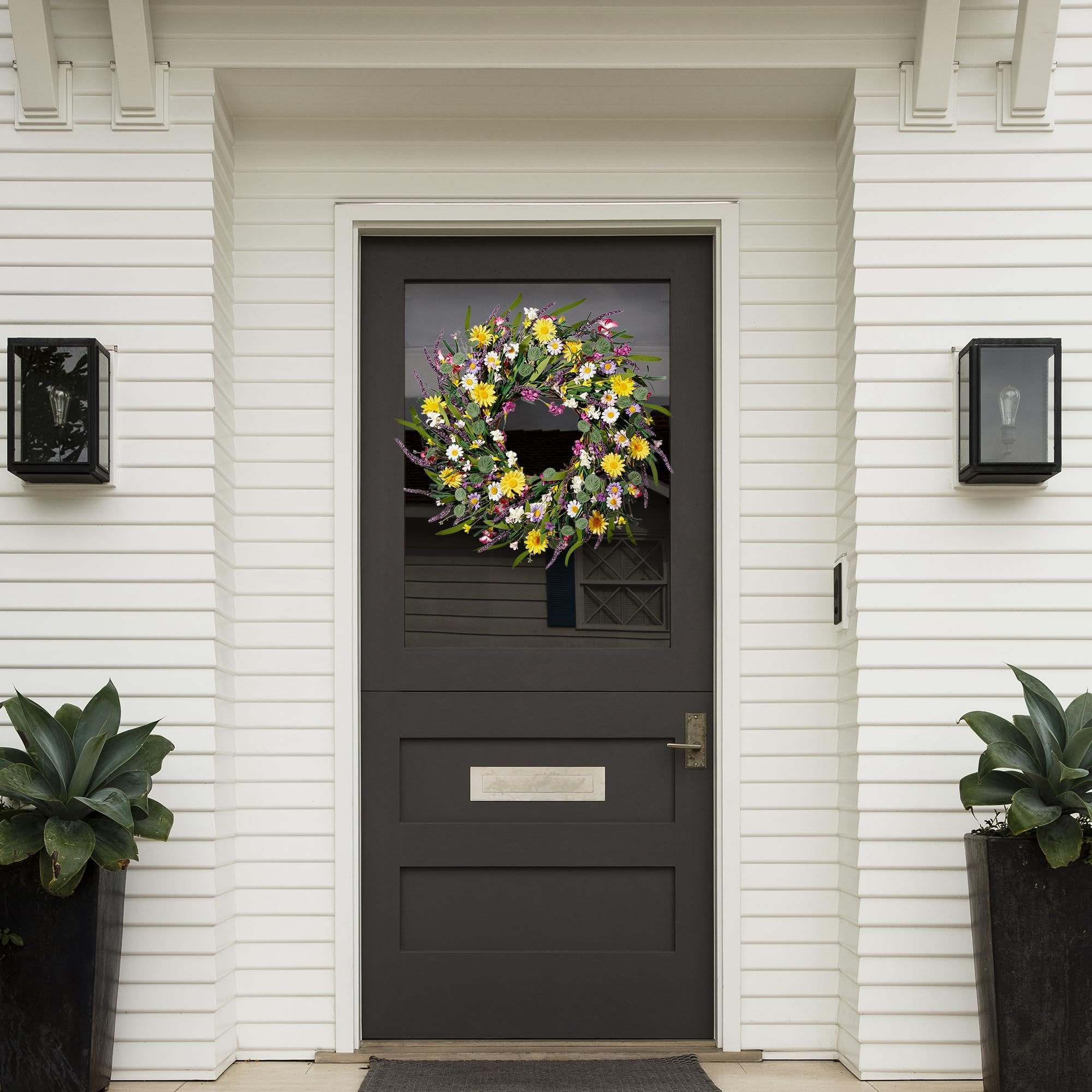 Artificial daisy and lavender wildflower wreath hanging on a dark front door with white siding.