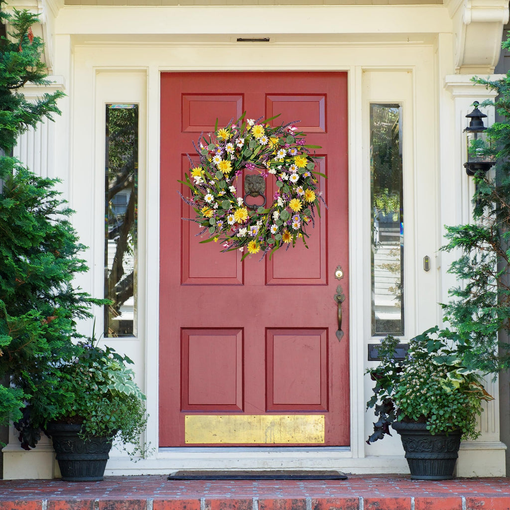 Artificial daisy and lavender wildflower wreath hanging on a red front door with decorative plants on either side.