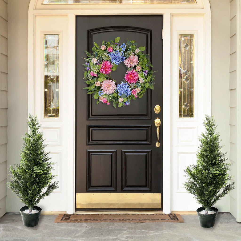 Floral wreath with pink, blue, and green flowers and leaves hanging on a black front door with two potted trees.