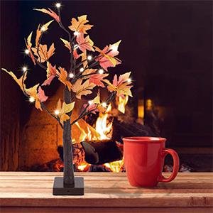 Artificial lighted maple tree with orange-red leaves and 24 LED lights, placed on a table next to a red mug with a fireplace in the background.
