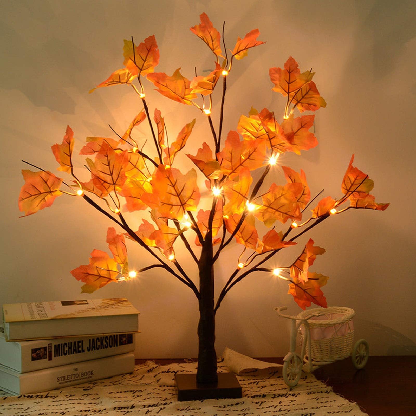 Lighted artificial maple tree with orange-red leaves and 24 LED lights, set on a table with books and a decorative tricycle.