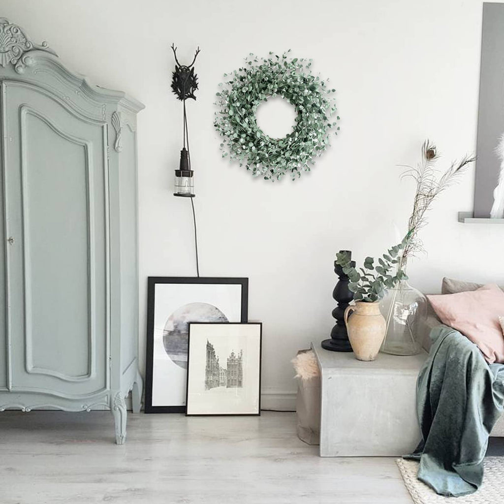 24 inch boxwood wreath with soft touch green leaves hanging on a white wall above framed art and a vintage cabinet.