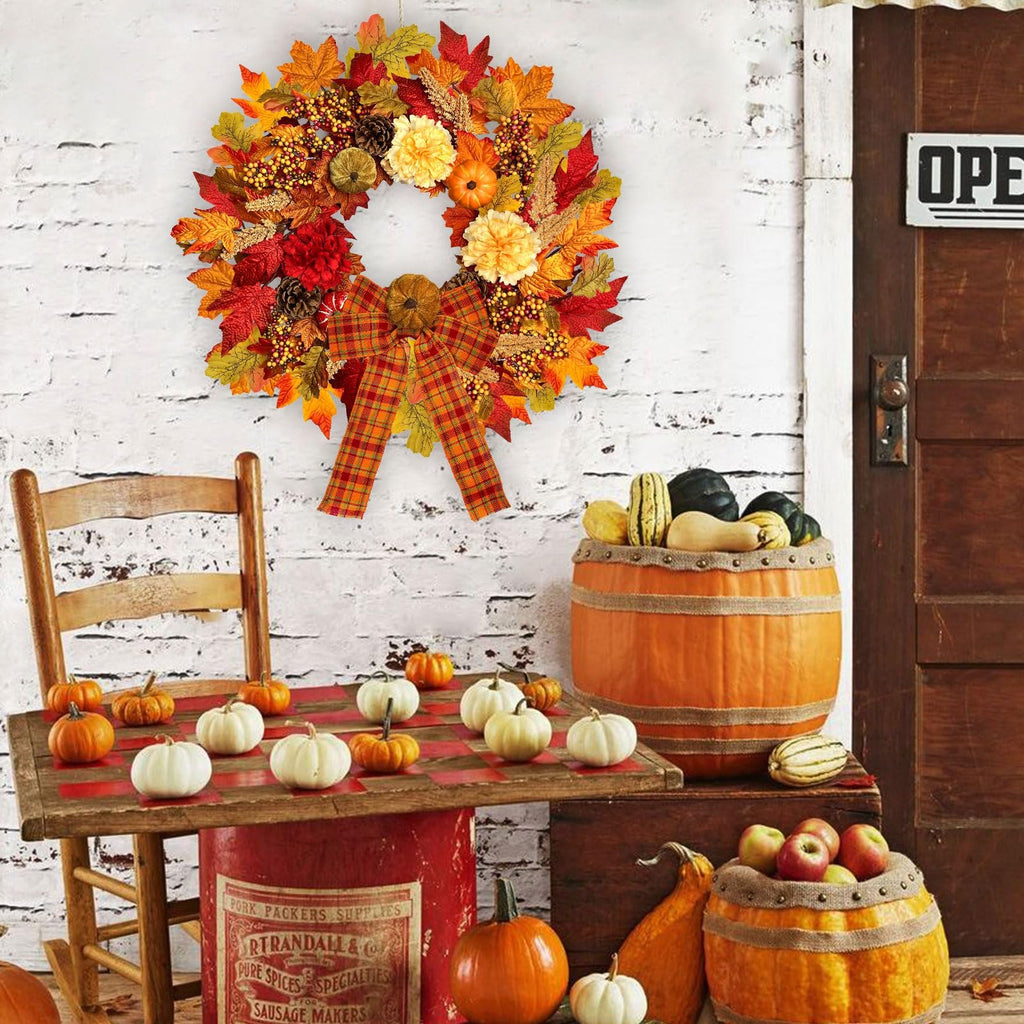 Fall wreath with pumpkins, berries, dahlias, and plaid bow hanging on a white brick wall above a table with pumpkins and gourds.