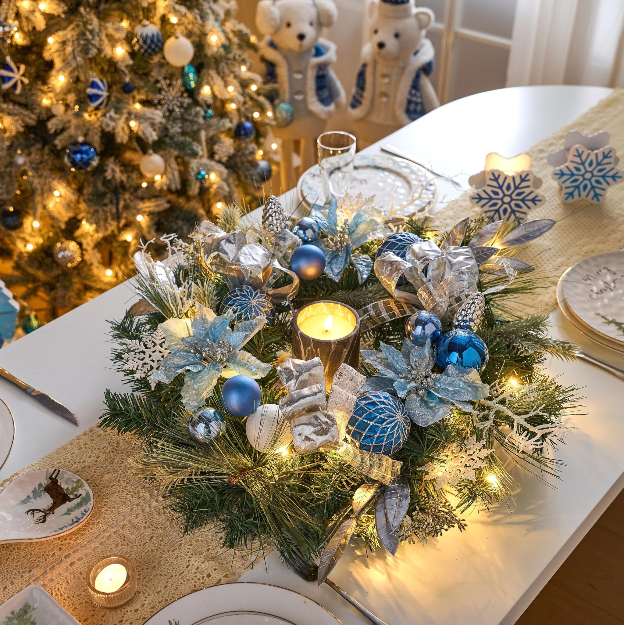 Christmas wreath with silver and blue ornaments, ribbons, and poinsettias on a table with a lit Christmas tree in the background.