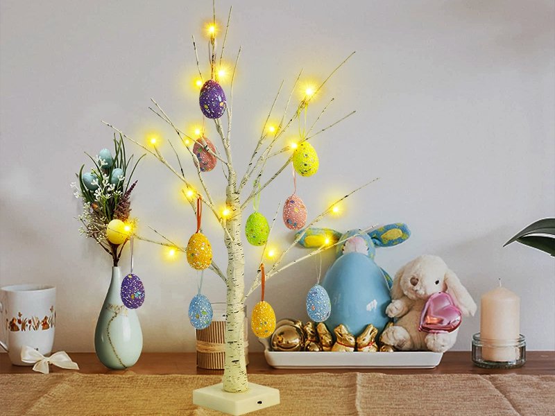 Pre-lit white birch Easter tree with colorful hanging egg ornaments and warm white LED lights, displayed with decorative bunnies and a candle.