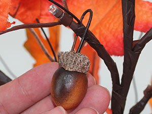 Close-up of a hand holding an artificial acorn ornament on a decorative maple tree with LED lights and orange leaves.