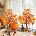 Two lighted maple trees with acorns and pine cones, set on a table with a pumpkin and craft supplies.