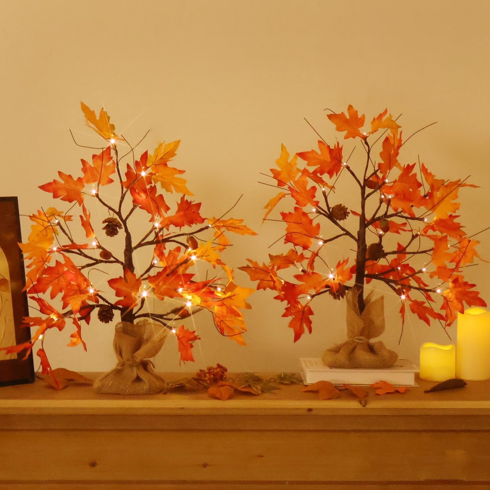 Two lighted maple trees with orange and yellow leaves and pine cones on a wooden shelf with candles.