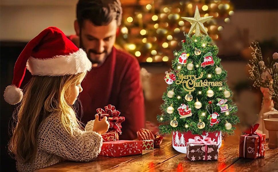 A young girl in a Santa hat and a man look at a decorated mini Christmas tree with gifts on a wooden table.