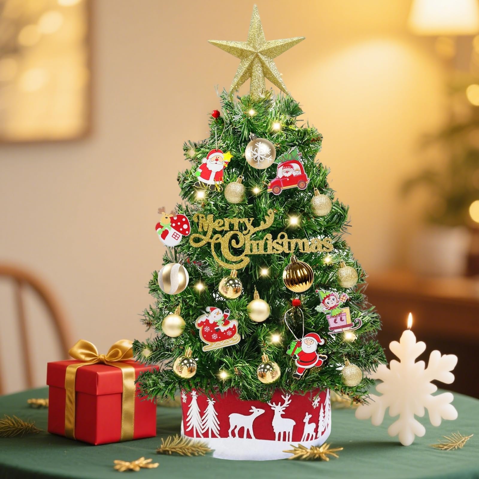 Miniature Christmas tree decorated with ornaments, lights, and a star topper, set on a table with a gift box and snowflake candle.