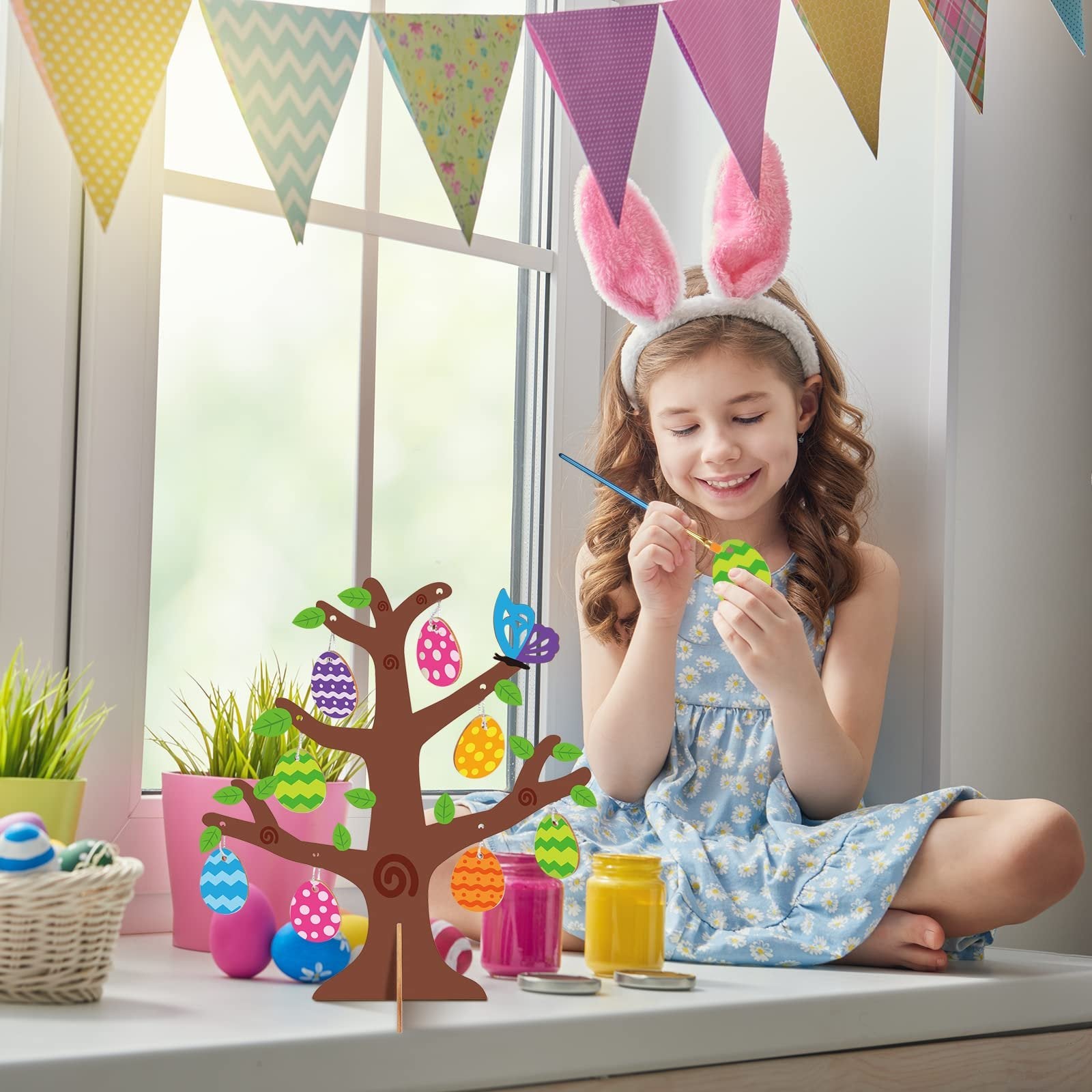 Girl painting Easter eggs on a wooden tree craft kit by a window with bunny ears