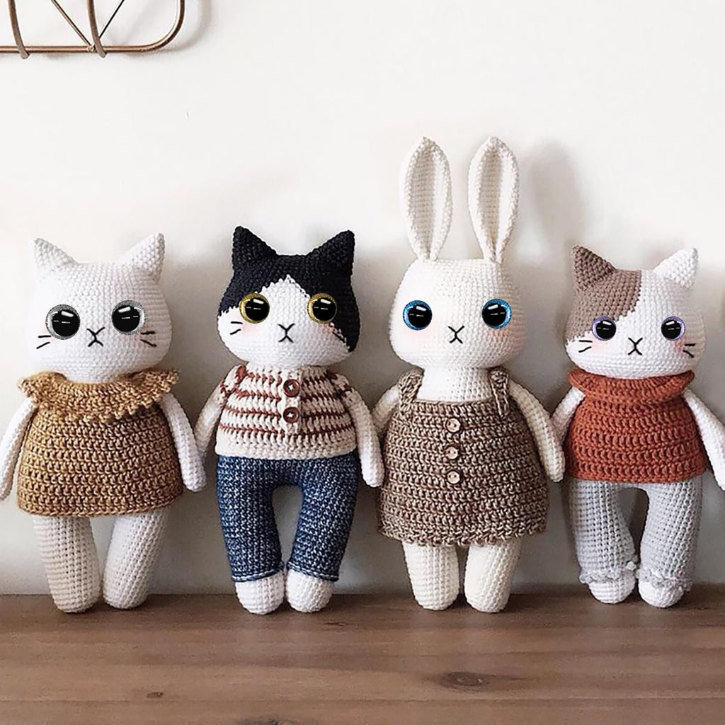 Four crocheted stuffed animals, two cats and one rabbit, standing side-by-side on a wooden shelf. The animals have large, colorful, glittery safety eyes and embroidered mouths and noses.