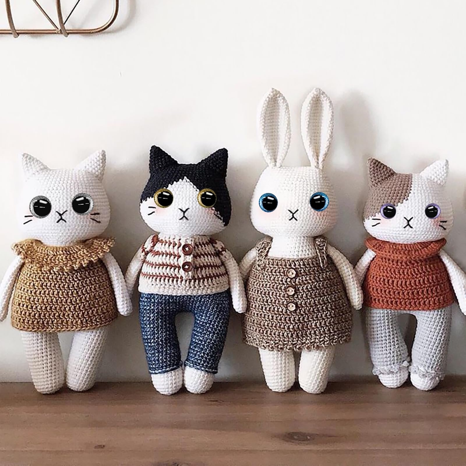 Four crocheted stuffed animals, two cats and one rabbit, standing side-by-side on a wooden shelf. The animals have large, colorful, glittery safety eyes and embroidered mouths and noses.