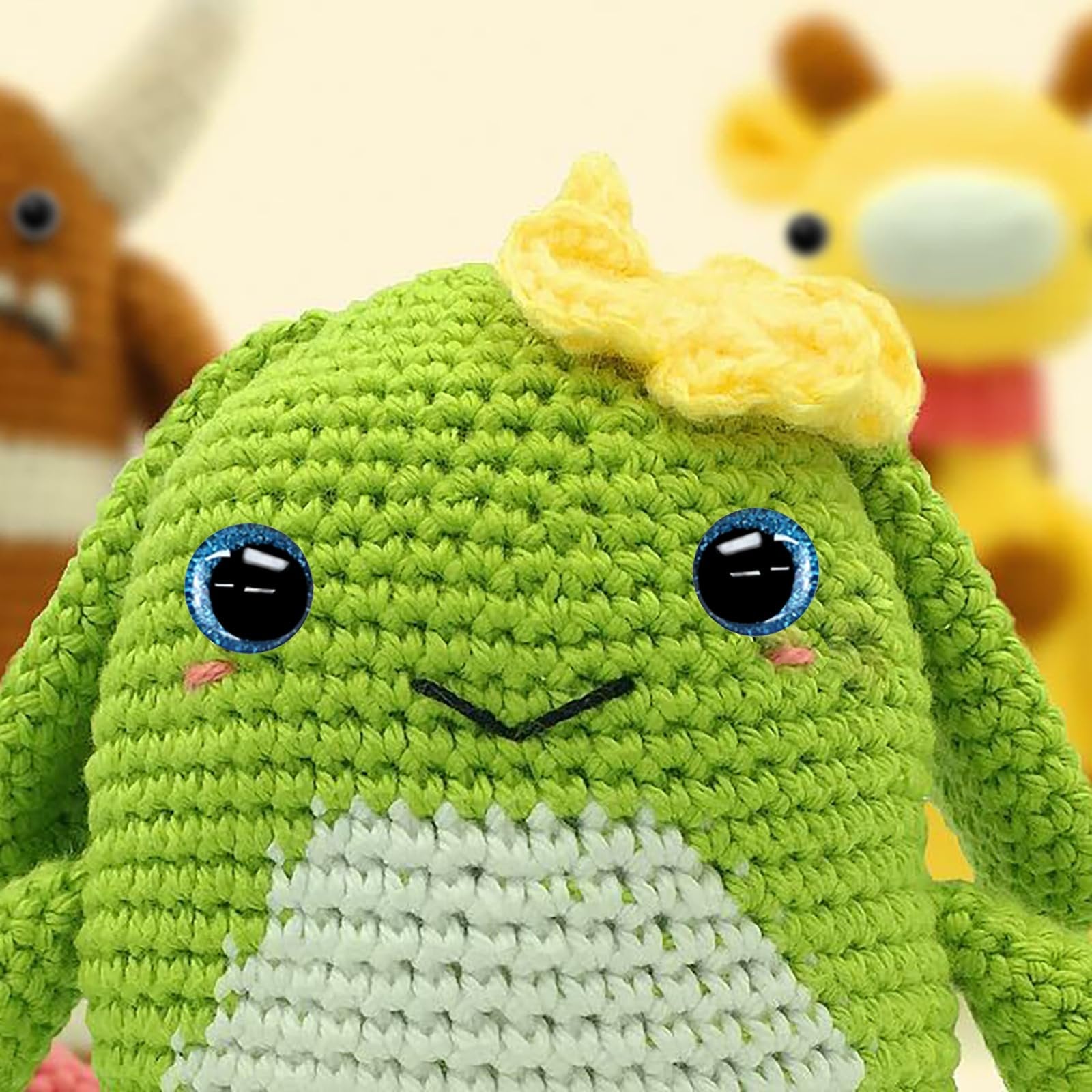 Close-up of a green crochet monster with blue glitter eyes and a yellow bow on its head, with other crochet toys blurred in the background.