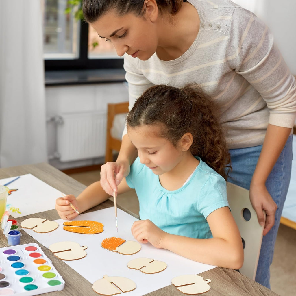 A woman helps a young girl paint unfinished wooden pumpkin craft pieces on a table with watercolors.
