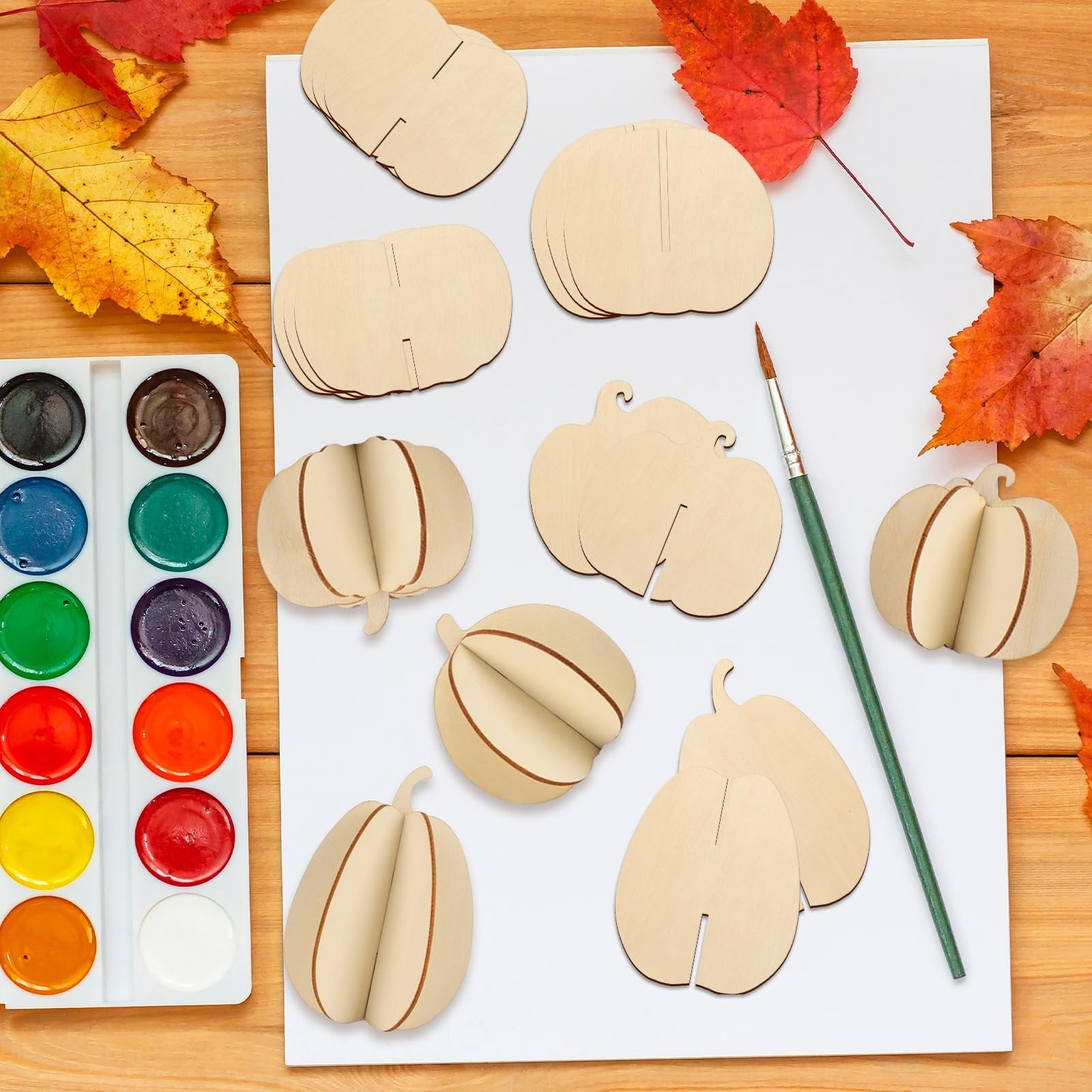 Unfinished wooden pumpkin craft pieces and a watercolor paint set with a brush on a wooden surface with autumn leaves.