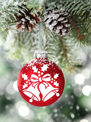 Red Christmas ornament with white stencil design of bells and holly hanging from a frosted pine branch with pinecones