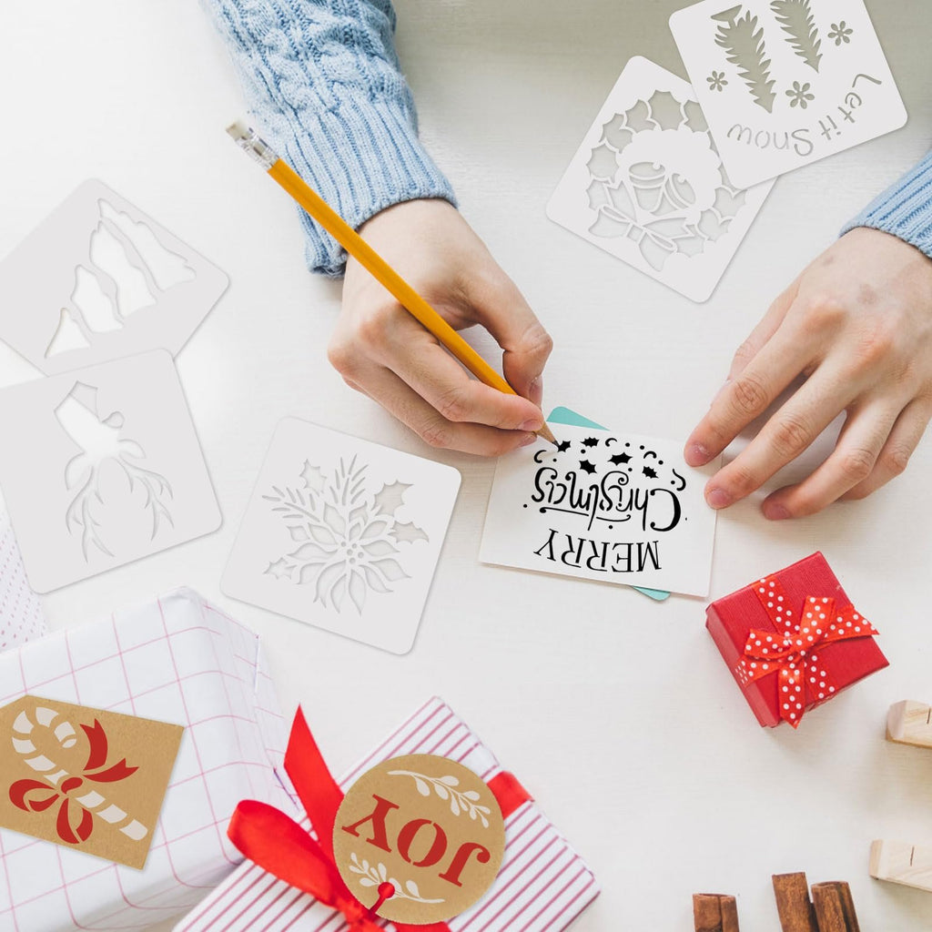 Hands using a pencil to trace a stencil onto a card that says Merry Christmas, surrounded by other Christmas stencils and wrapped gifts.