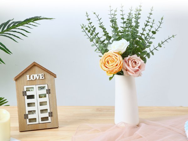 Artificial eucalyptus stems with roses in a vase next to a wooden house-shaped frame with the word LOVE