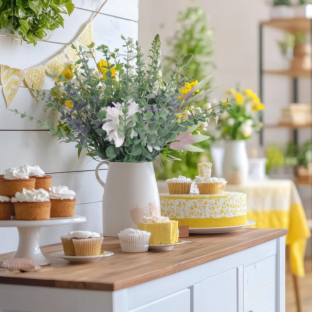 Artificial eucalyptus stems in a white pitcher with cupcakes and a cake on a wooden cabinet
