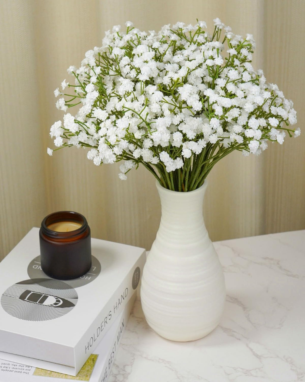 Bouquet of 24 white artificial baby's breath flowers in a white vase on a table with books and a candle.