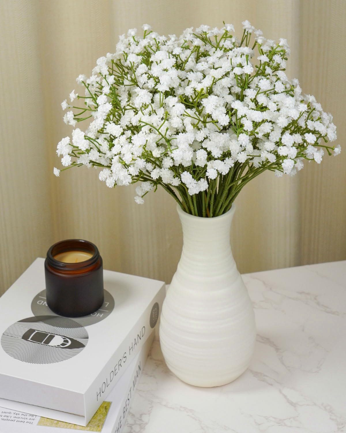 Bouquet of 24 white artificial baby's breath flowers in a white vase on a table with books and a candle.