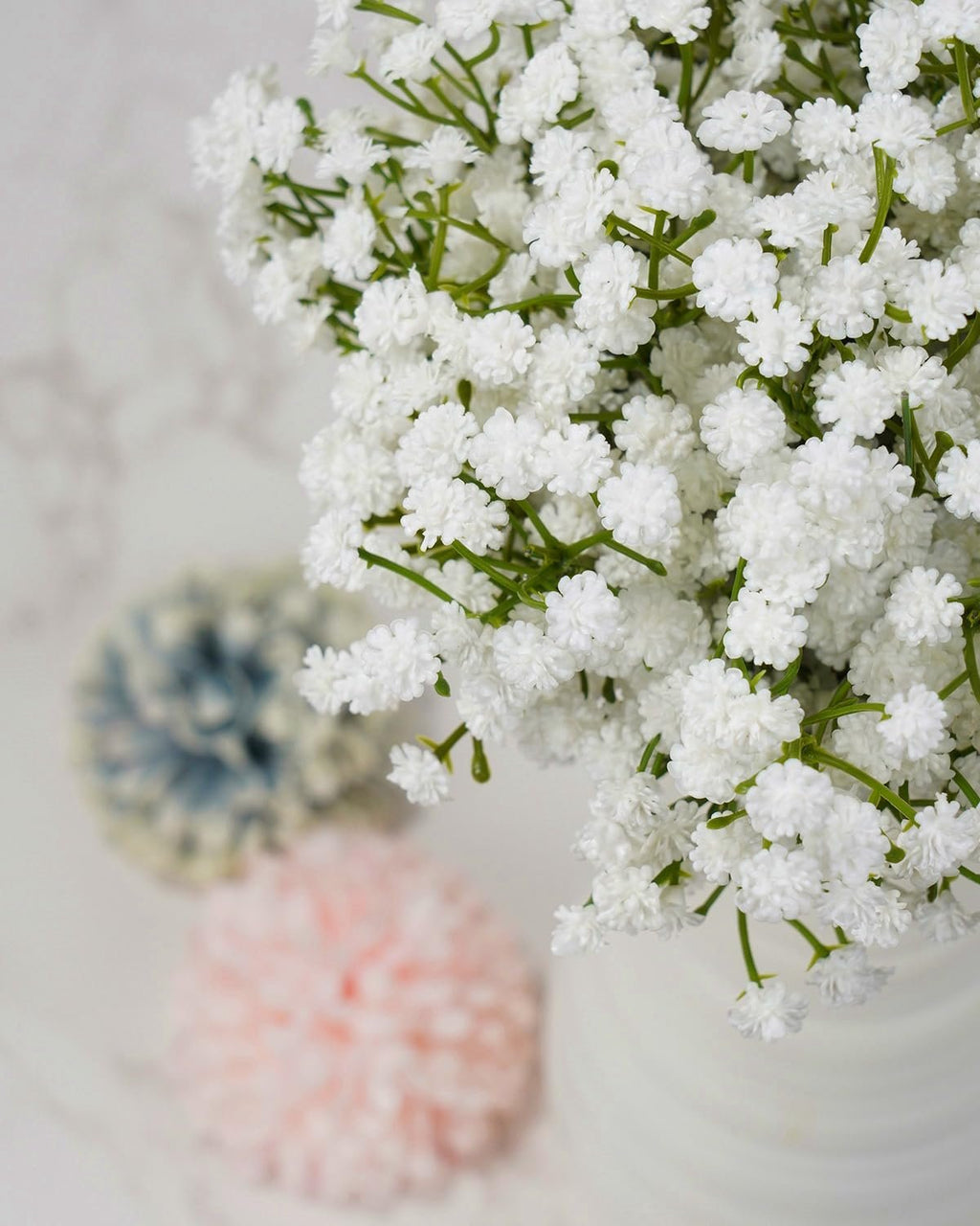 Close-up of white artificial baby's breath flowers with green stems, blurred pink and blue pom-poms in background.
