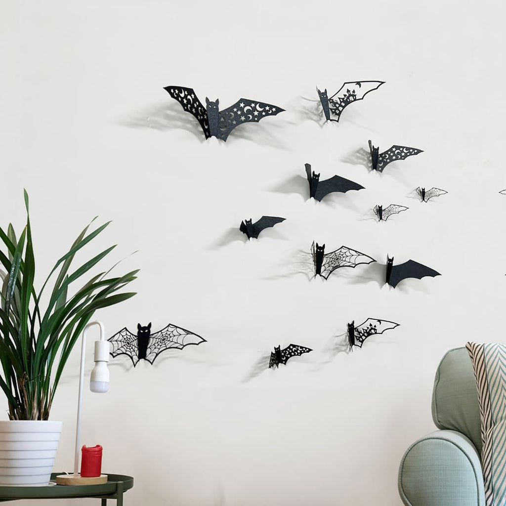 Black 3D Halloween bat wall stickers in various sizes arranged on a white wall, with a potted plant and chair visible.