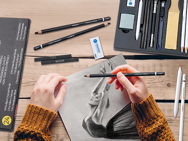 Hands drawing a portrait with a charcoal pencil set, showing pencils, erasers, and blending stumps on a wooden table.