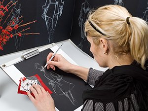 Woman sketching fashion designs on black paper with charcoal pencils, surrounded by art supplies and sketches.