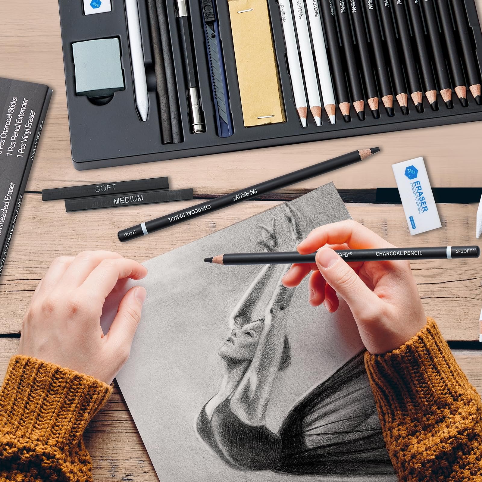Hands drawing a portrait with a charcoal pencil set, including pencils, erasers, and charcoal sticks.