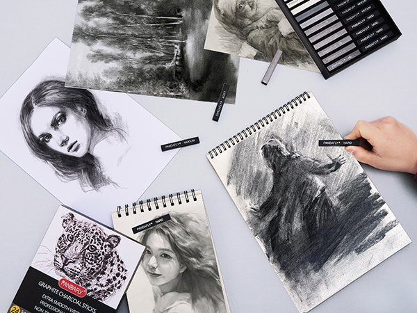 Drawing set with charcoal sticks and sketchpads featuring portraits and abstract art. A hand holds a charcoal stick.