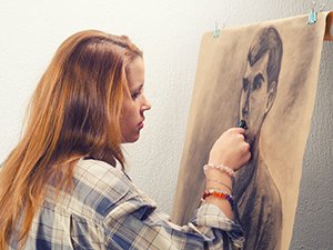 Young woman drawing a portrait on brown paper with charcoal sticks, viewed from the side