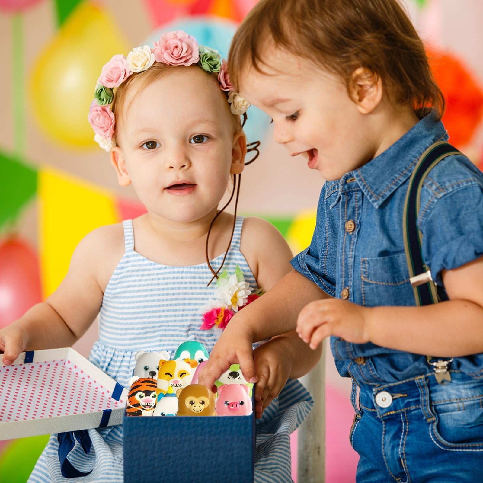 Two toddlers looking into a box of assorted slow-rising squishy animal toys, with balloons in the background.