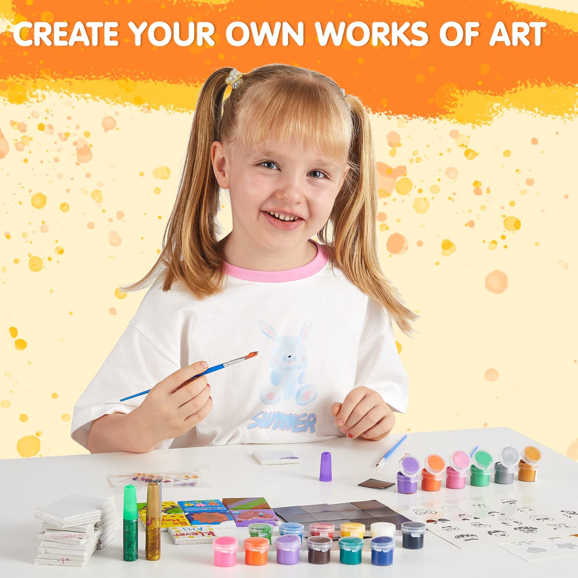 Smiling young girl painting magnetic tiles with colorful paints and glitter glue, art kit supplies laid out on table.